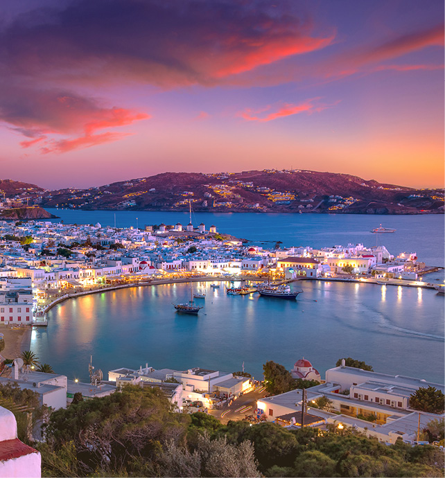 An evening view of Mykonos, with boats seen in the harbour, hills in the background, and twinkling lights in the buildings. 