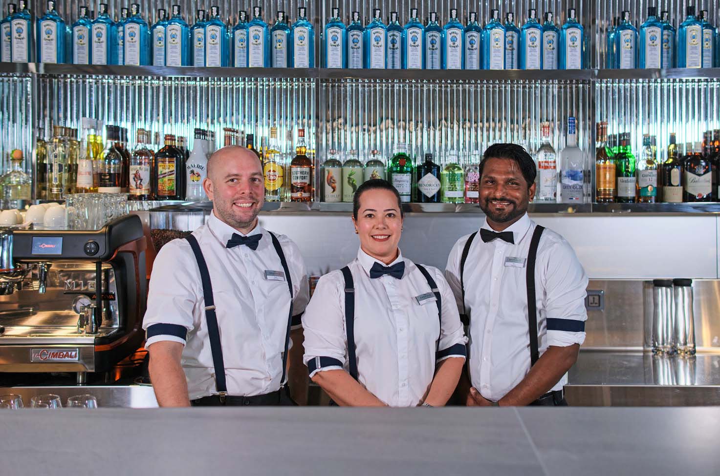 Three bar staff posing behind a fully stocked bar on a cruise